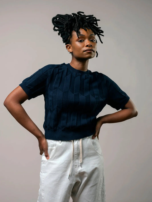 A model with short black dreadlocks is wearing a dark navy blue short-sleeve knit top with a textured geometric pleated pattern, paired with white drawstring pants, against a plain light gray background.