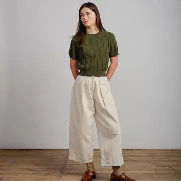A woman stands in a studio wearing an olive green short-sleeve knit top and off-white wide-leg cropped pants, paired with brown shoes, against a light gray wall and wood floor.