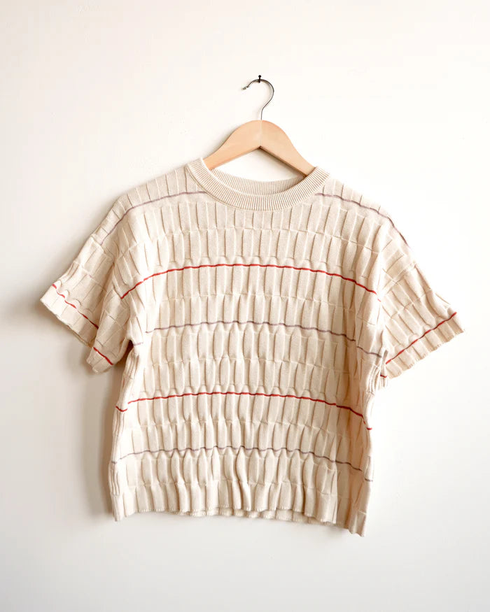 A cream-colored short-sleeve knit top with horizontal pleated texture and thin red stripe accents, hanging on a wooden hanger against a plain off-white wall.