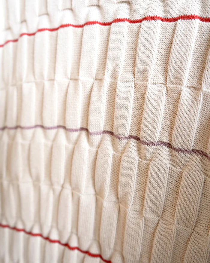 Close-up of a cream-colored textured knit fabric with a basketweave pattern, featuring thin horizontal red and purple stripes.