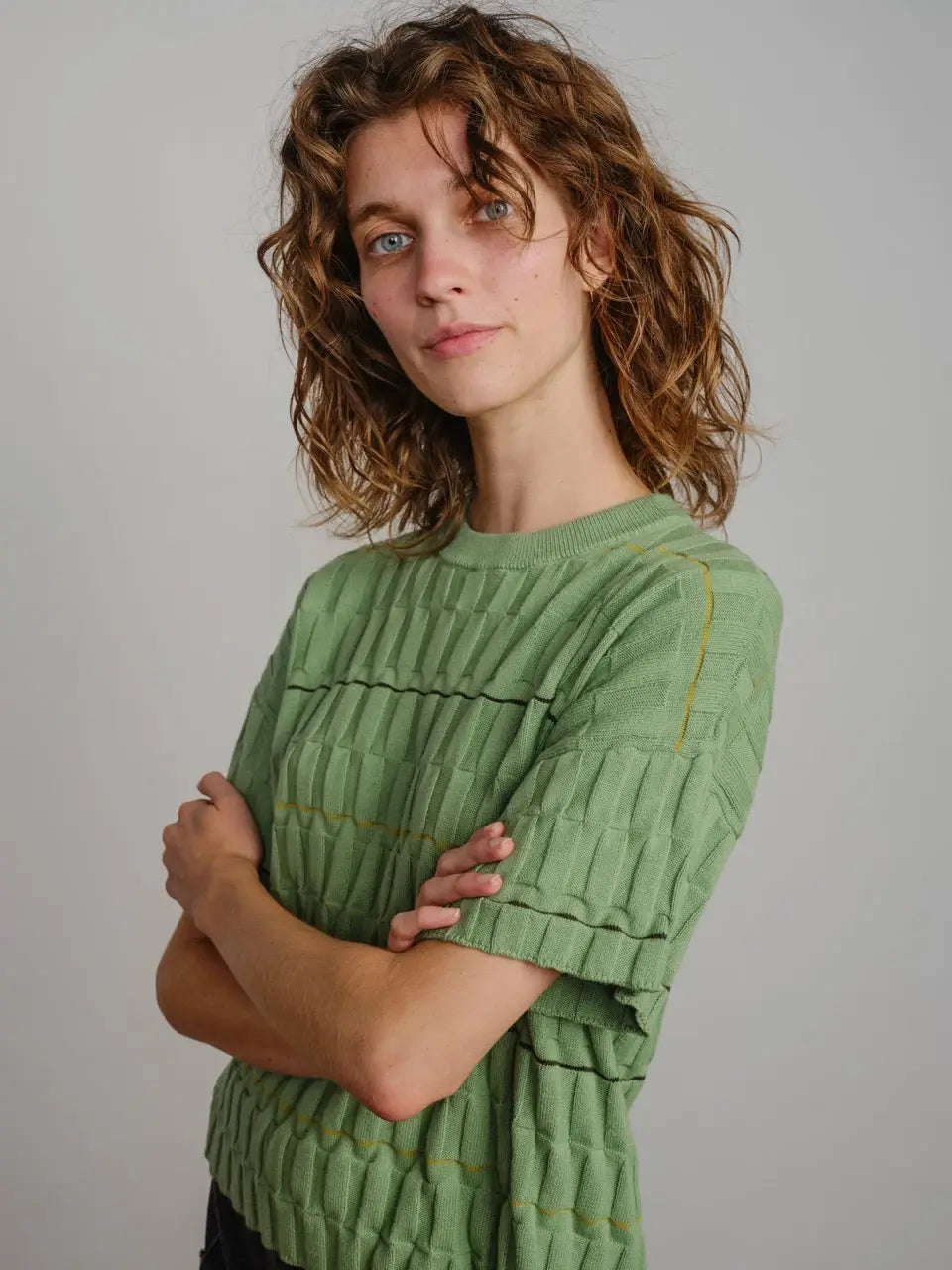Woman sitting on a wooden chair wearing a green textured sweater and light-colored pants against a plain wall.