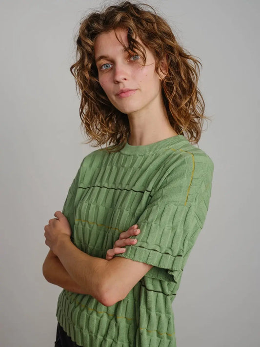 Woman sitting on a wooden chair wearing a green textured sweater and light-colored pants against a plain wall.