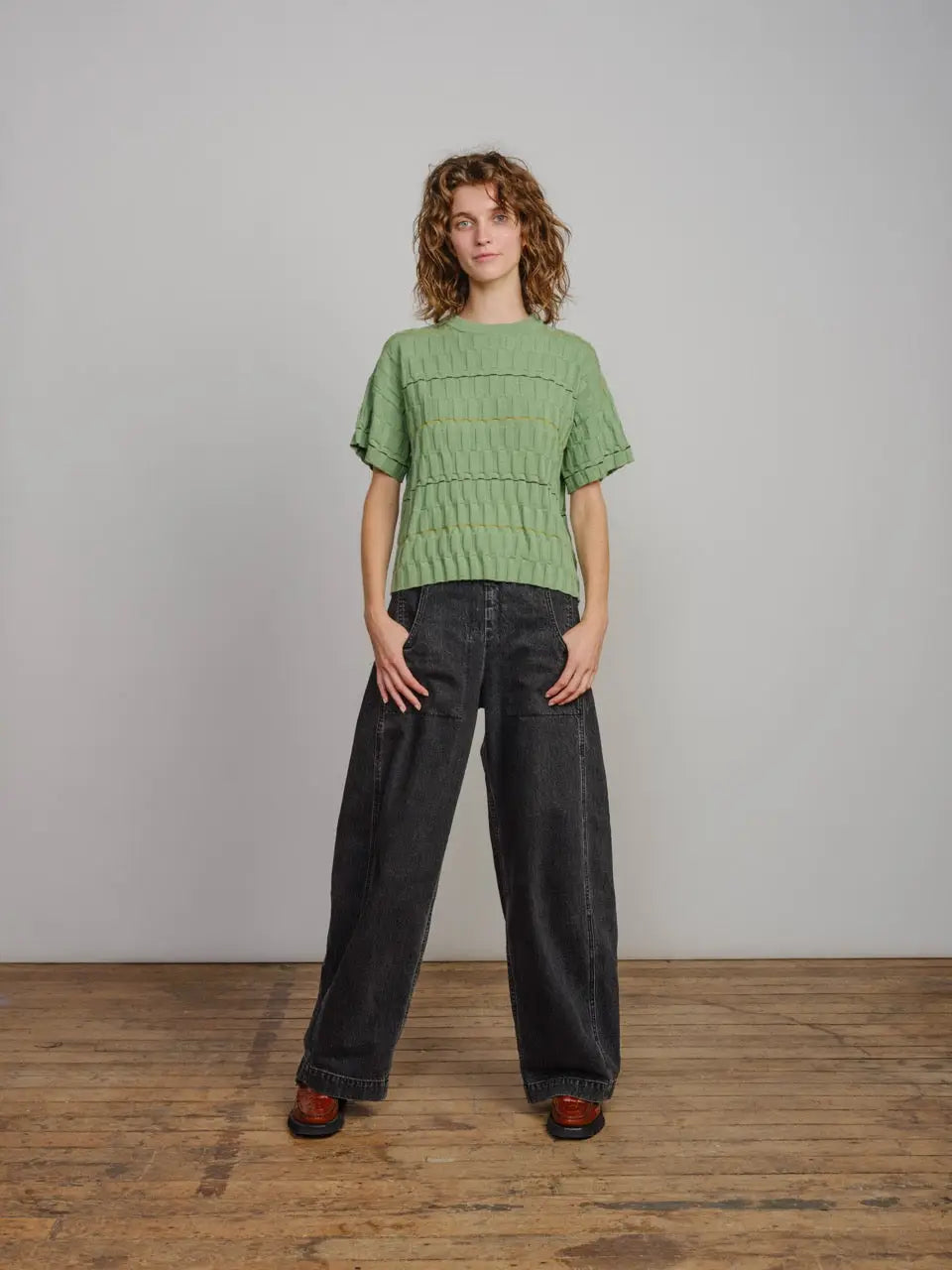 Woman standing wearing a green textured sweater and dark-colored pants against a plain wall.