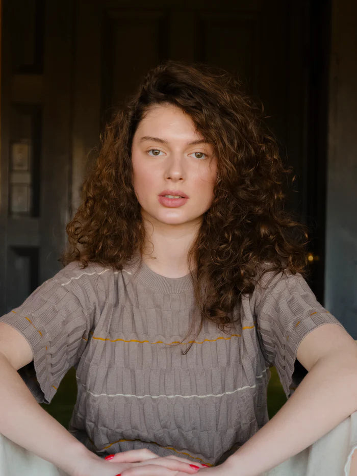 A woman with curly brown hair wearing a textured taupe-gray short-sleeve top with thin yellow and white horizontal stripe details, posed in front of a dark wooden door.