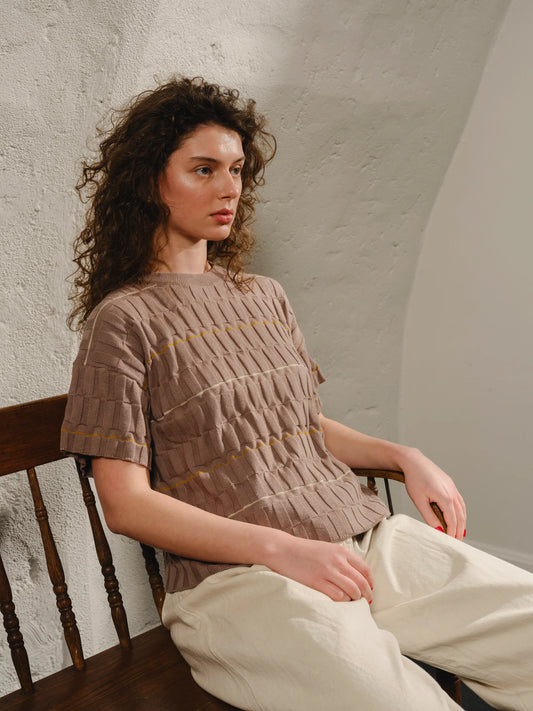 Woman sitting on a wooden chair wearing a brown textured sweater and light-colored pants against a plain wall.