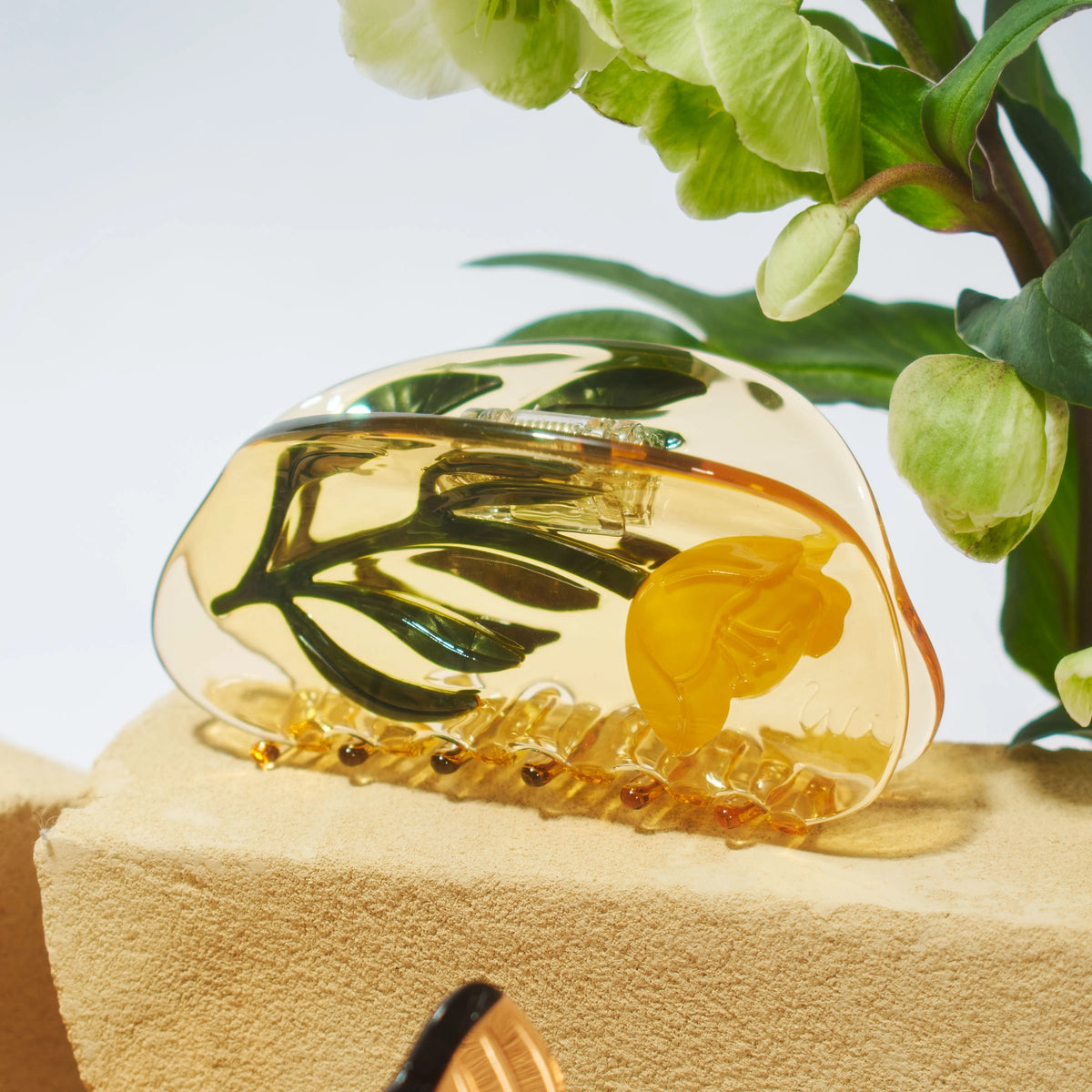 Gold decorative claw hair clip with leaf design on a beige surface with green leaves in the background