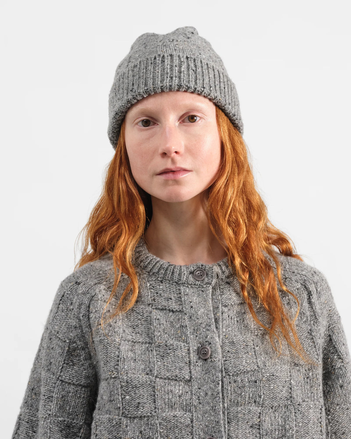 A woman with long red hair wearing a gray knit beanie and a matching gray textured knit cardigan, photographed against a plain light gray studio background.