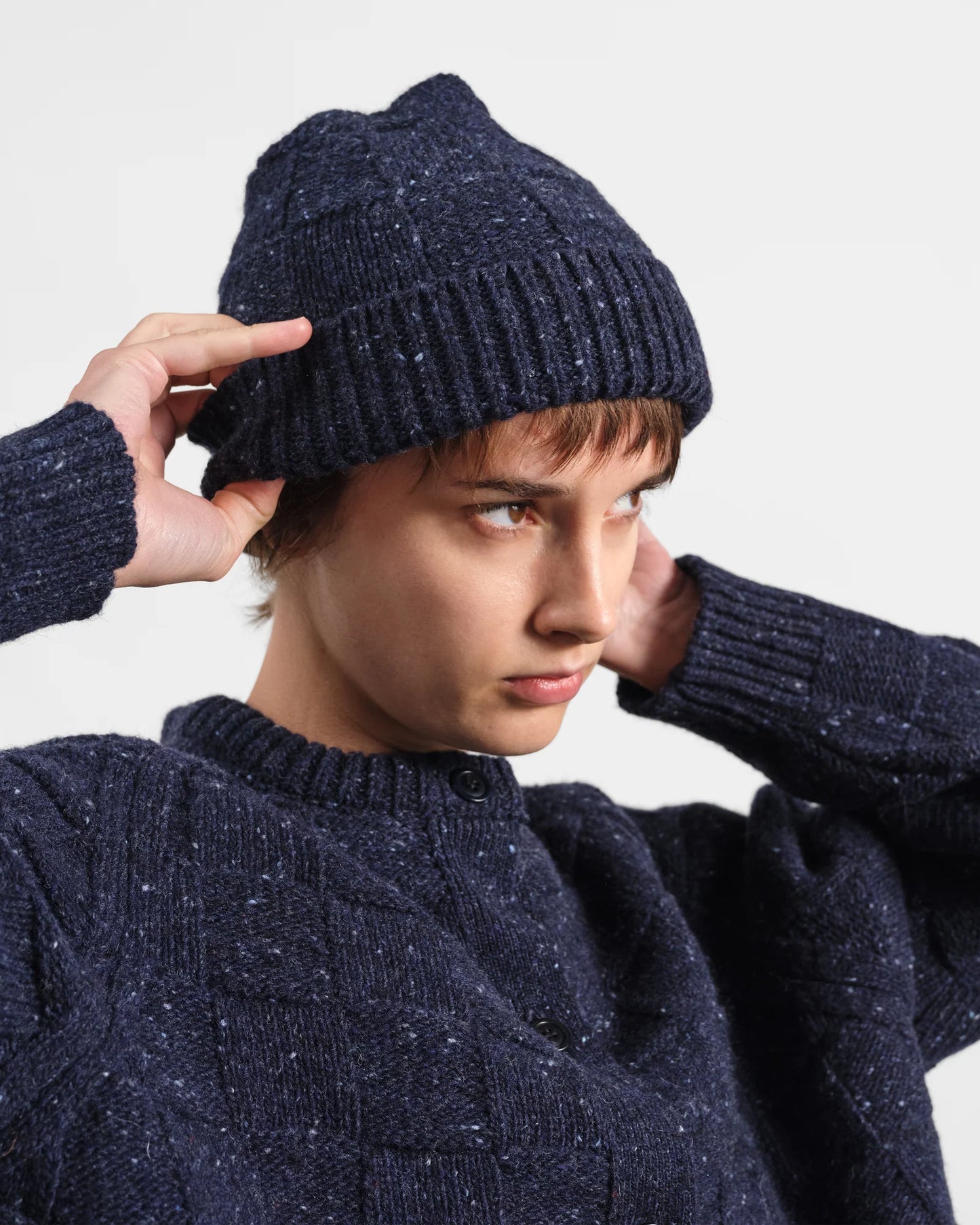 A person wearing a dark navy knit beanie and matching navy sweater, adjusting the beanie with one hand against a plain light gray background.