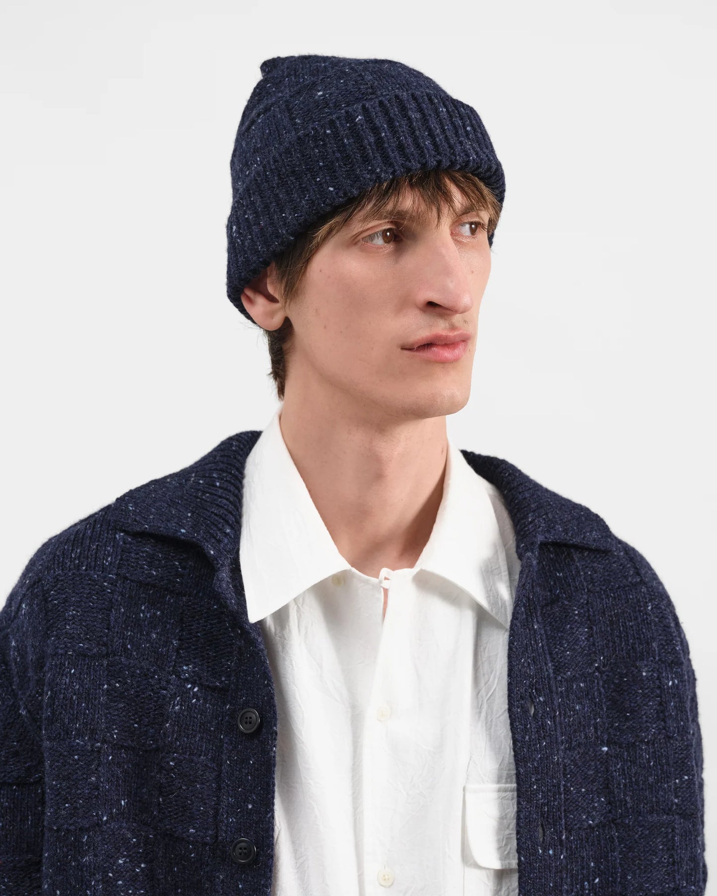 A man wearing a dark navy blue knit beanie, a white collared shirt, and a dark navy blue textured knit cardigan, photographed against a clean light gray studio background.