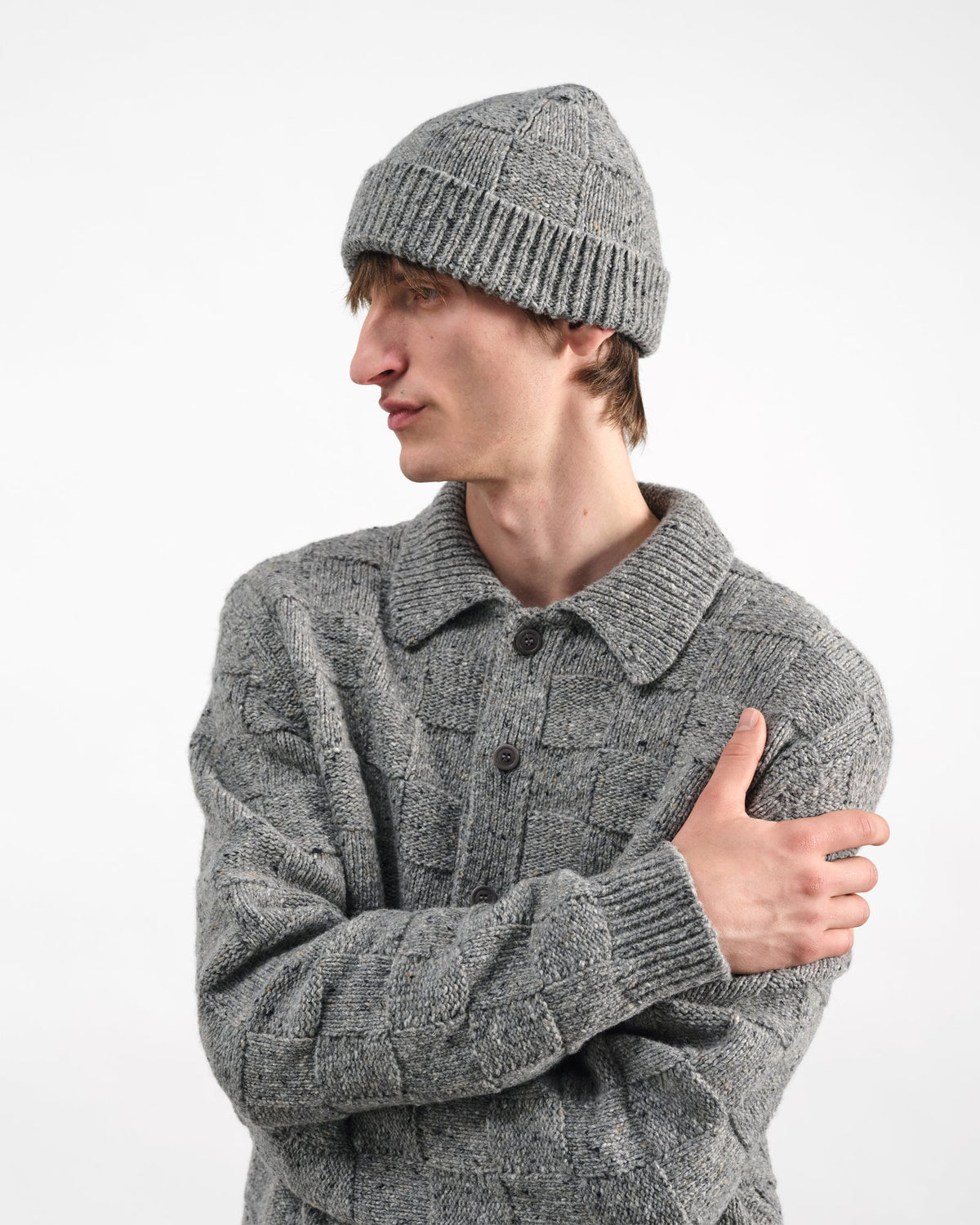 A man wearing a gray textured knit beanie and matching gray cable-knit button-up sweater, posed against a plain white studio background.