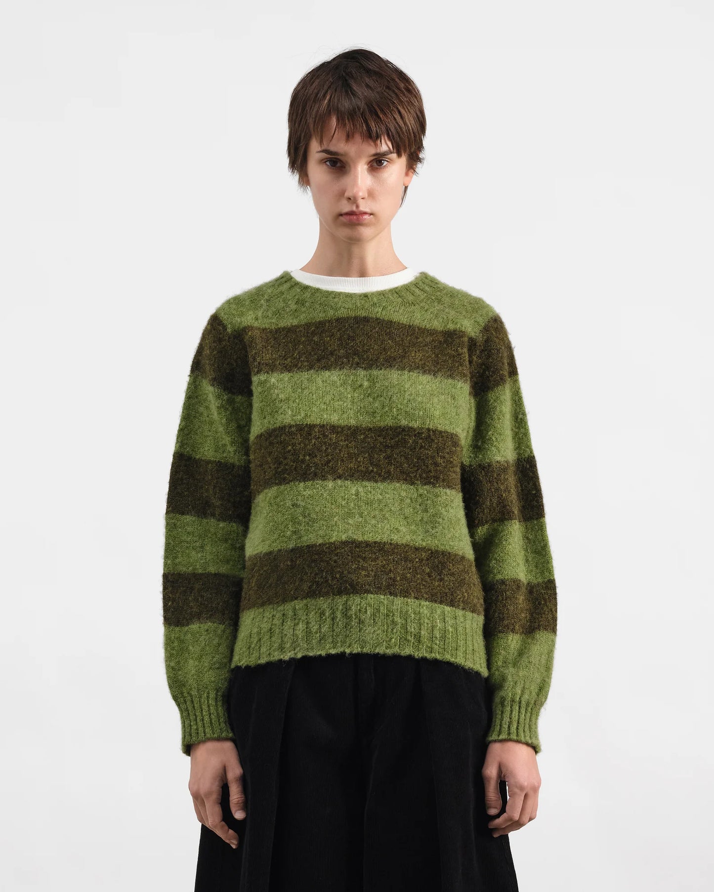 A model wearing a fuzzy, striped knit sweater with horizontal bands of olive green and dark brown, paired with a white crewneck undershirt and black wide-leg trousers, shot against a plain white studio background.
