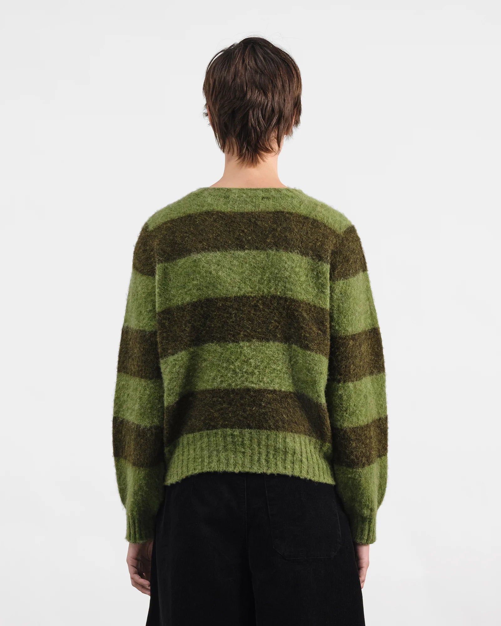 A person shown from the back wearing a fuzzy knit sweater with horizontal olive green and dark brown stripes, paired with black pants, against a plain white studio background.