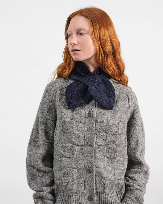 A model with red hair wearing a gray cable-knit cardigan sweater and a dark navy blue knitted bow tie-neck scarf, photographed against a plain light gray background.