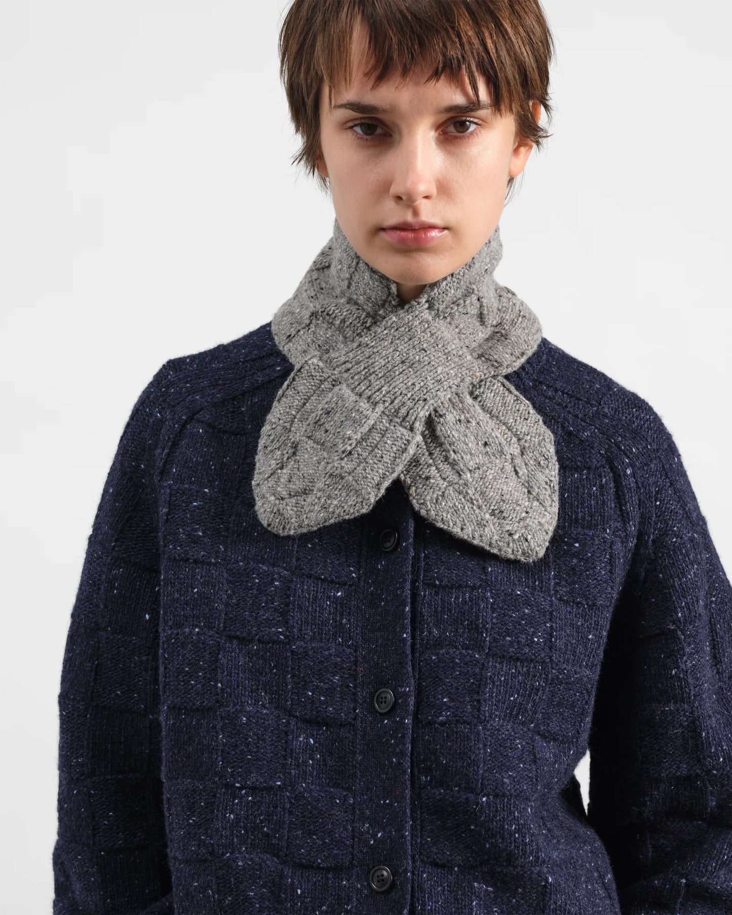 A model wearing a chunky gray cable-knit neck warmer scarf and a navy blue textured tweed button-up cardigan jacket, shot against a plain light gray background.
