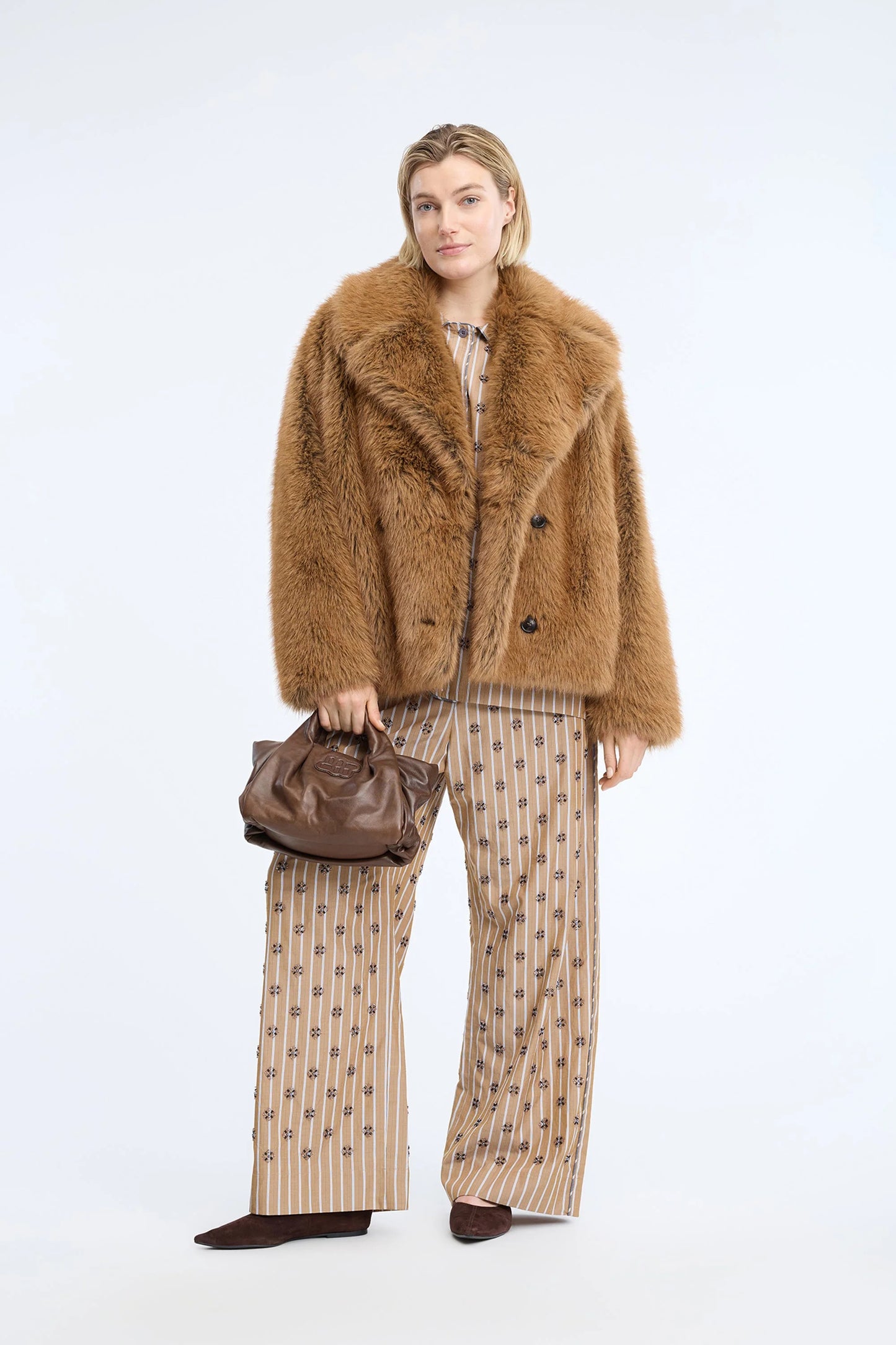 A model stands against a light gray studio background wearing a brown shaggy faux-fur jacket over a patterned top, wide-leg beige pants with a small floral print, and holding a brown leather handbag.