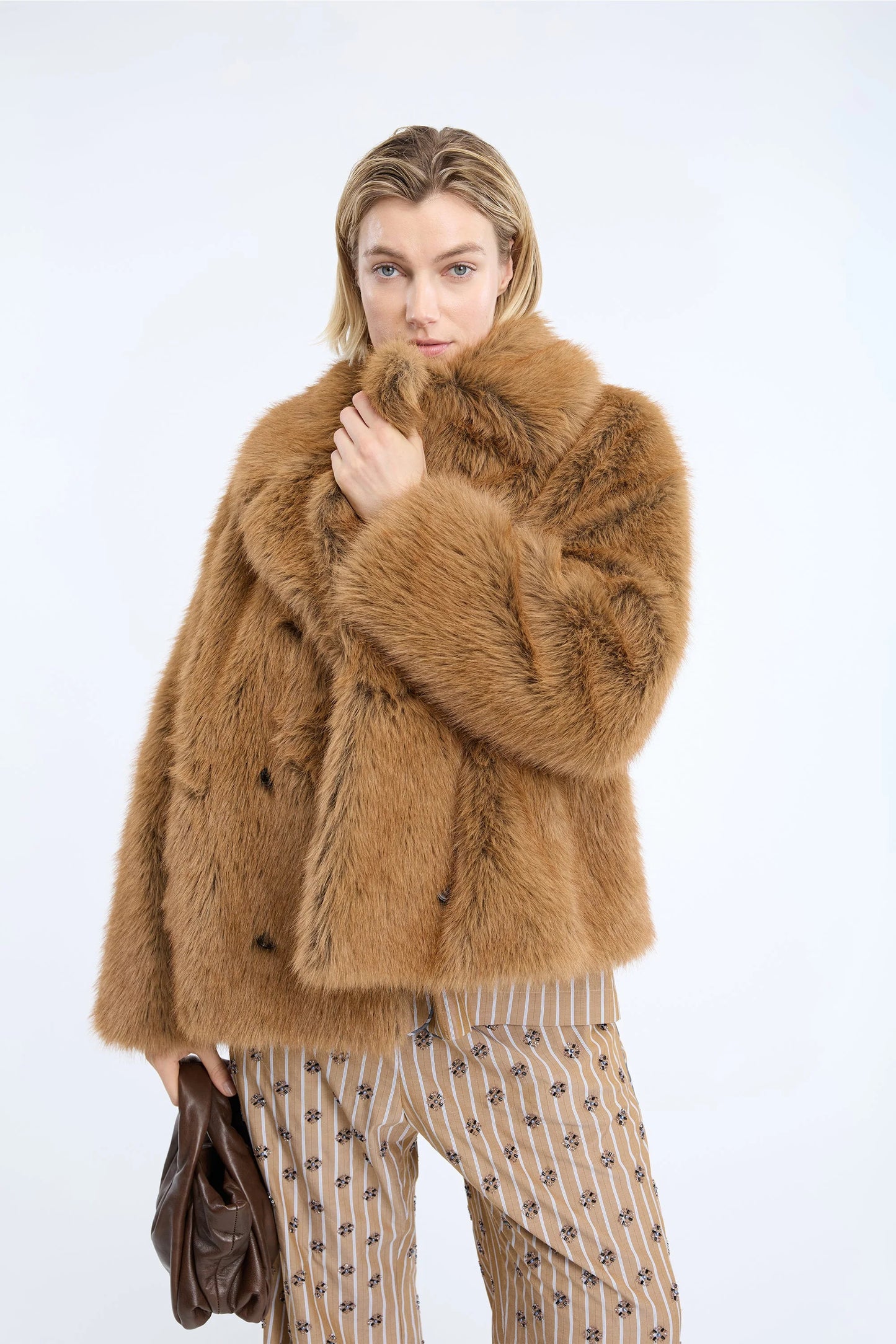 A model wearing a plush, double-breasted camel-brown fur coat with a high collar, paired with beige and brown patterned trousers, holding a dark brown leather bag against a plain white studio background.