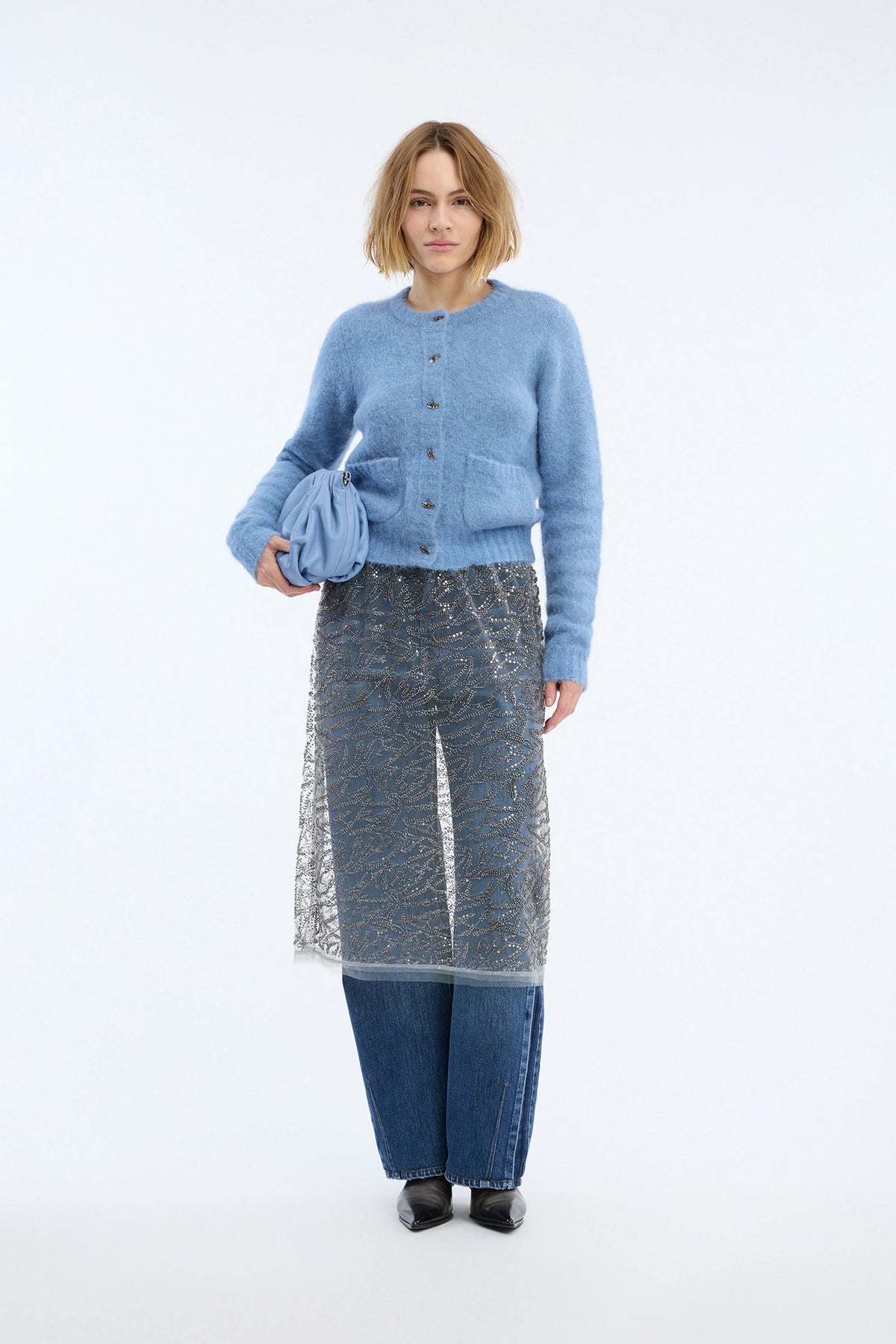 A model stands against a plain white studio background wearing a light blue knit cardigan, a sheer patterned gray-and-blue overlay skirt, wide-leg blue jeans, and black shoes, while holding a matching blue fabric bag.