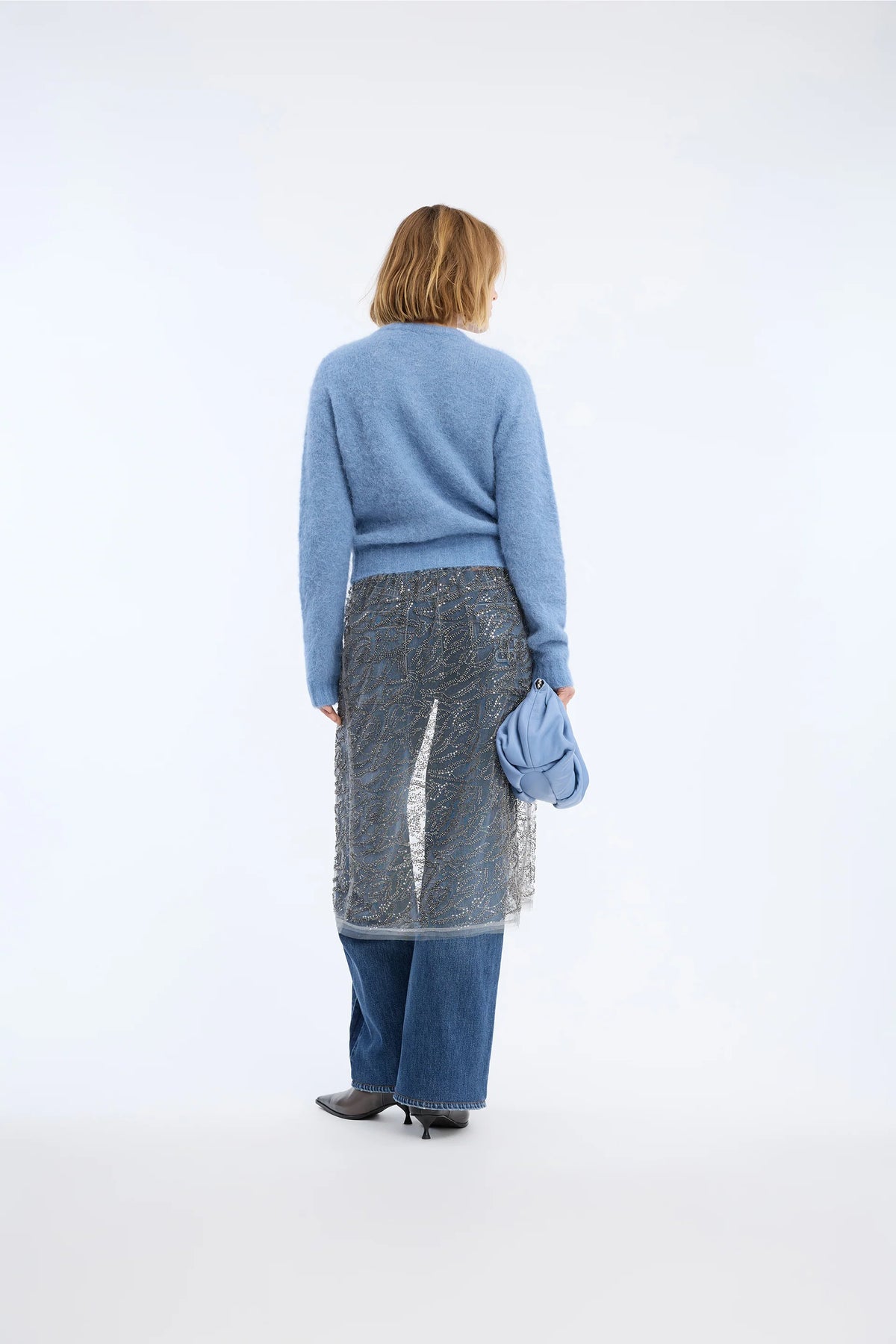 A model is shown from behind wearing a light blue knit sweater, a sheer gray patterned midi skirt, wide-leg blue jeans, and black pointed shoes, holding a matching light blue clutch bag against a plain white studio background.