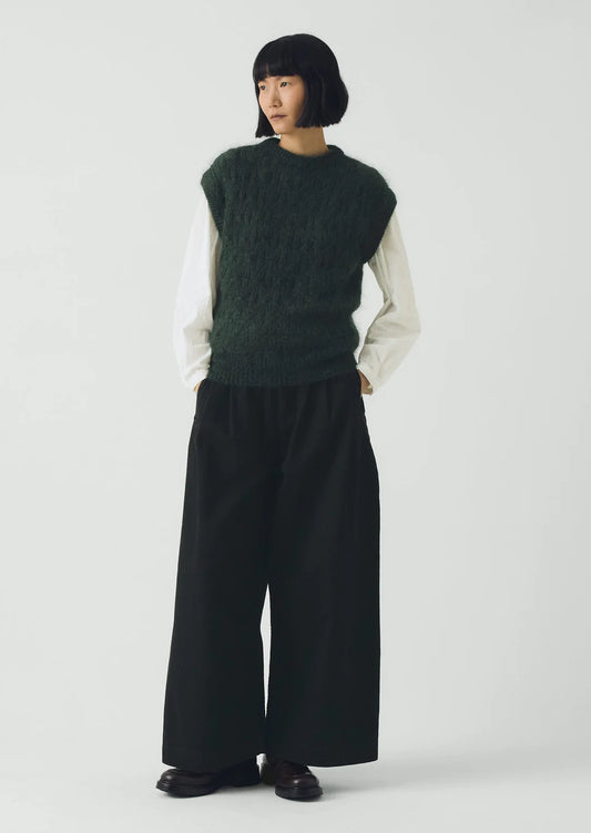 Person wearing a green knitted vest over a white shirt with black wide-leg pants on a white background