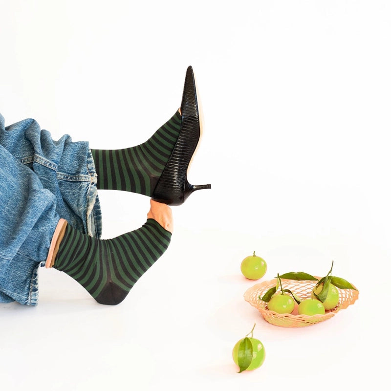 Person wearing black high-heeled boots with green striped socks, lying on a white surface with green fruits and a basket.