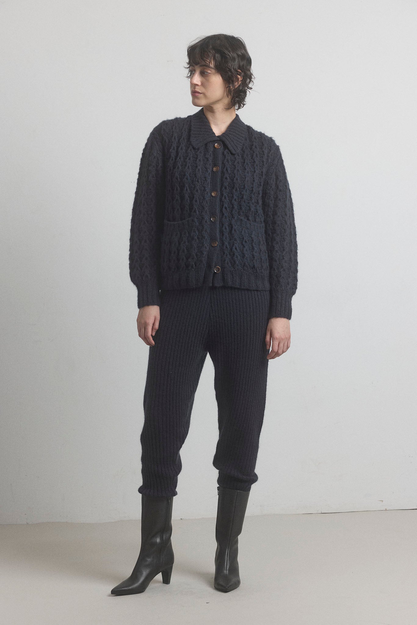 A person standing wearing merino cashmere ribbed joggers with a drawstring waistband, in a dark color, paired with a button-up shirt and boots.