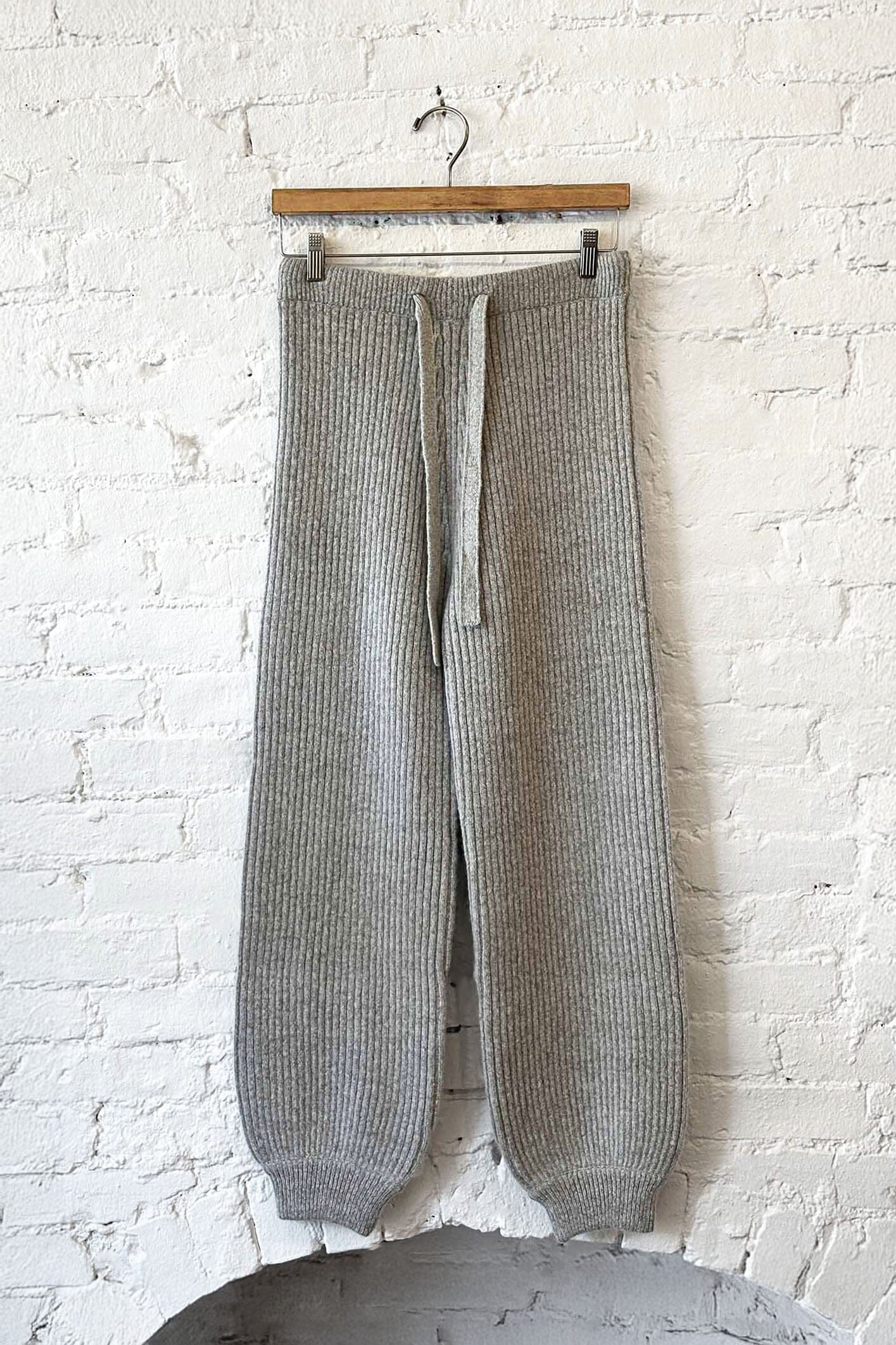 Gray knitted pants hanging on a wooden hanger against a white brick wall.