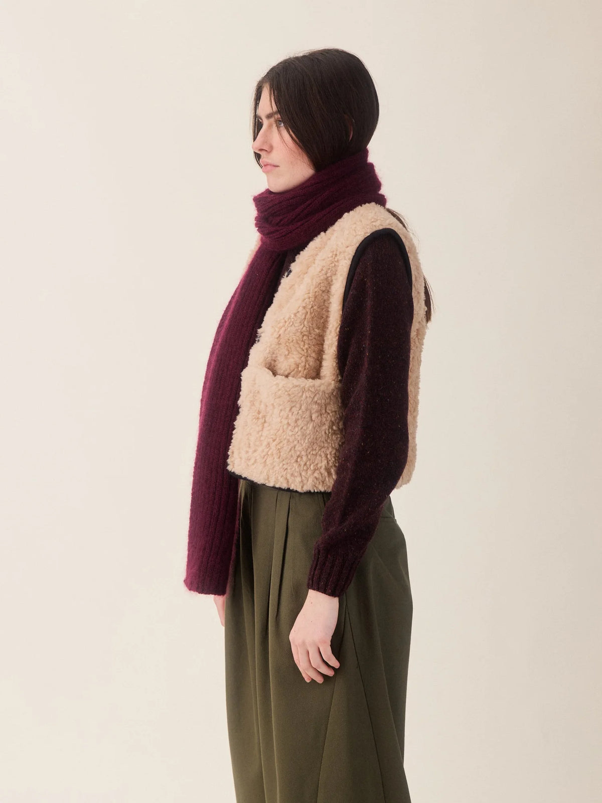 A model shown in profile wearing a beige sherpa vest over a dark burgundy knit sweater, paired with an olive green pants and a matching burgundy knit scarf, against a plain light beige background.