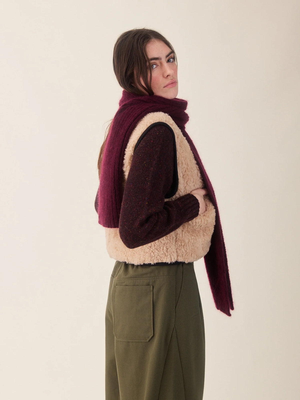 A woman wearing a cream sherpa vest over a dark burgundy knit sweater, paired with a matching deep burgundy knit scarf and olive green trousers, photographed against a solid light beige background.