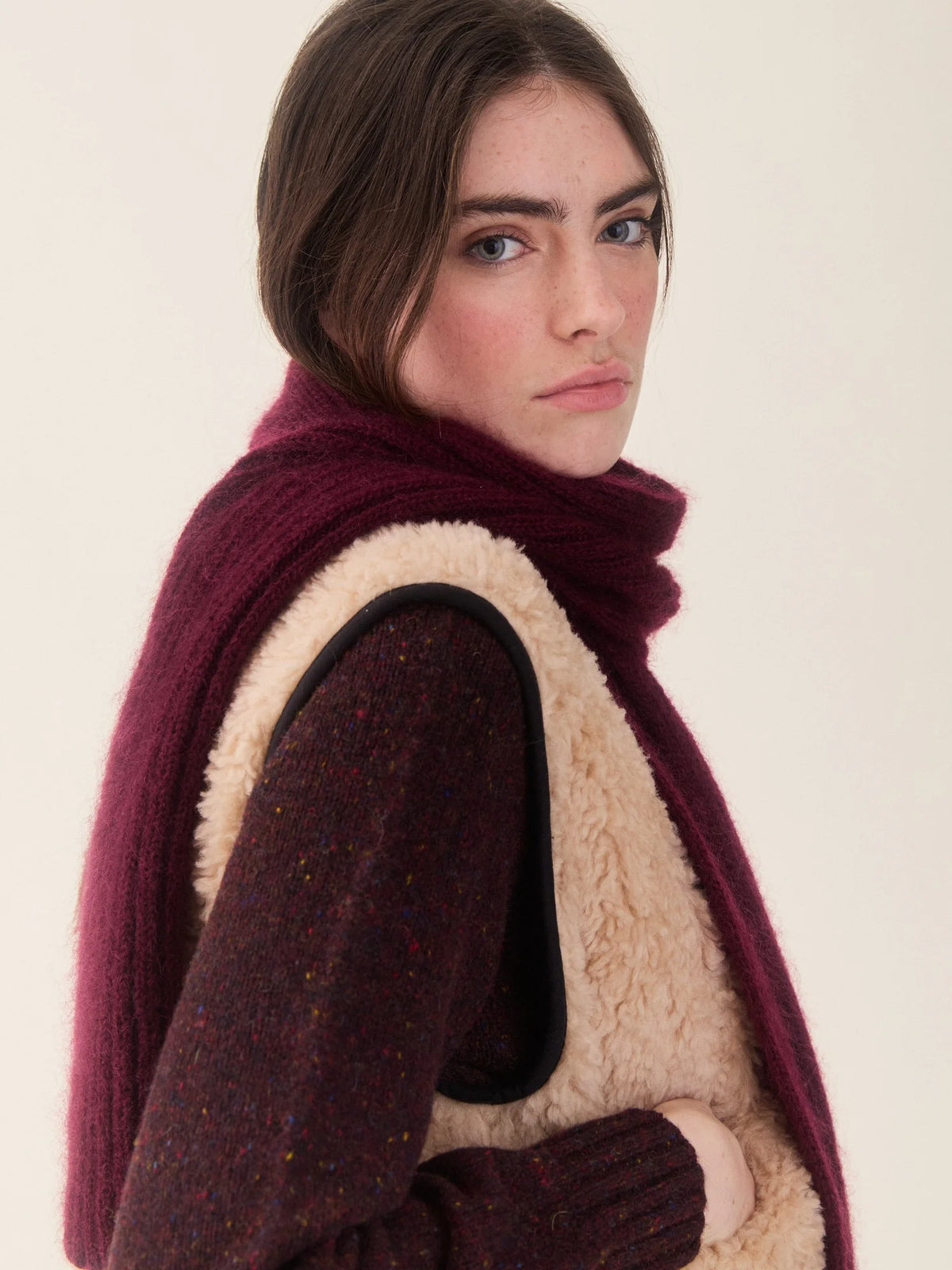 A woman wearing a deep burgundy knit scarf and a matching burgundy knit sweater vest with a cream sherpa lining, photographed against a plain off-white background.