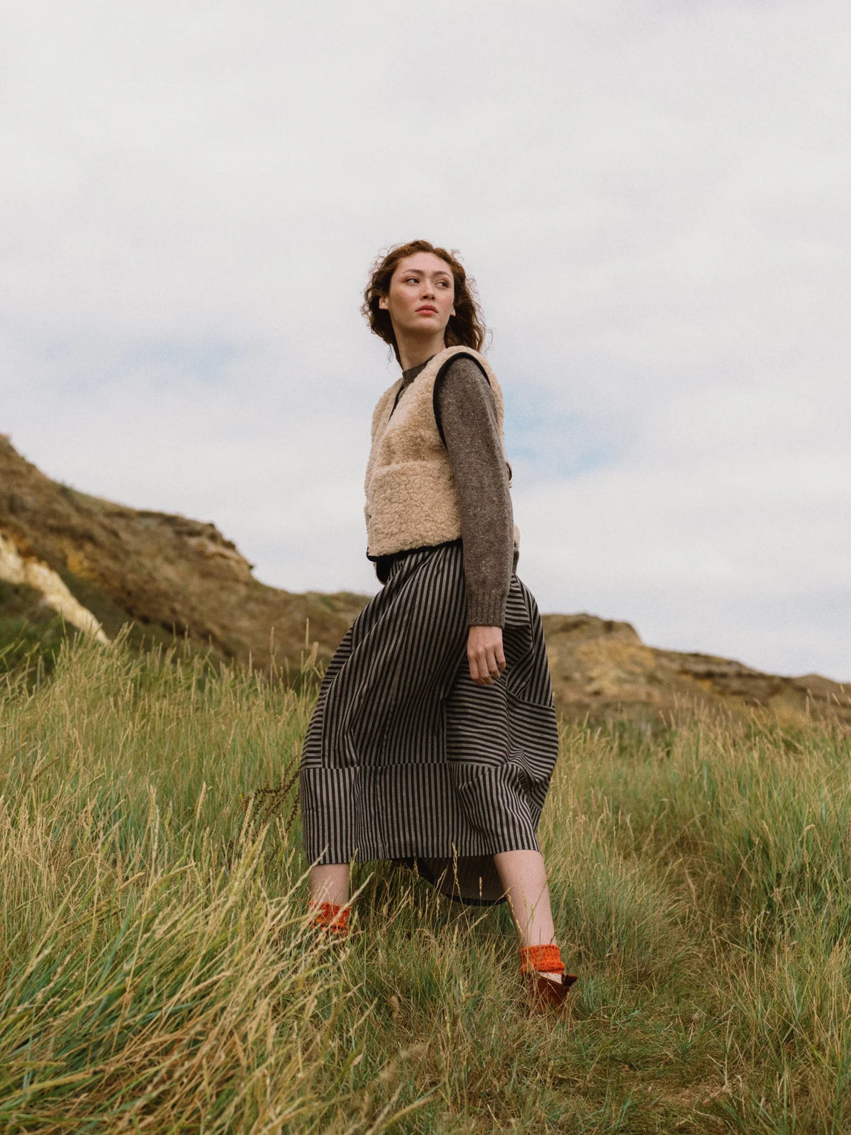 A woman stands in a grassy outdoor landscape wearing a beige fuzzy vest over a brown long-sleeve top and a dark striped midi skirt, with orange shoes.