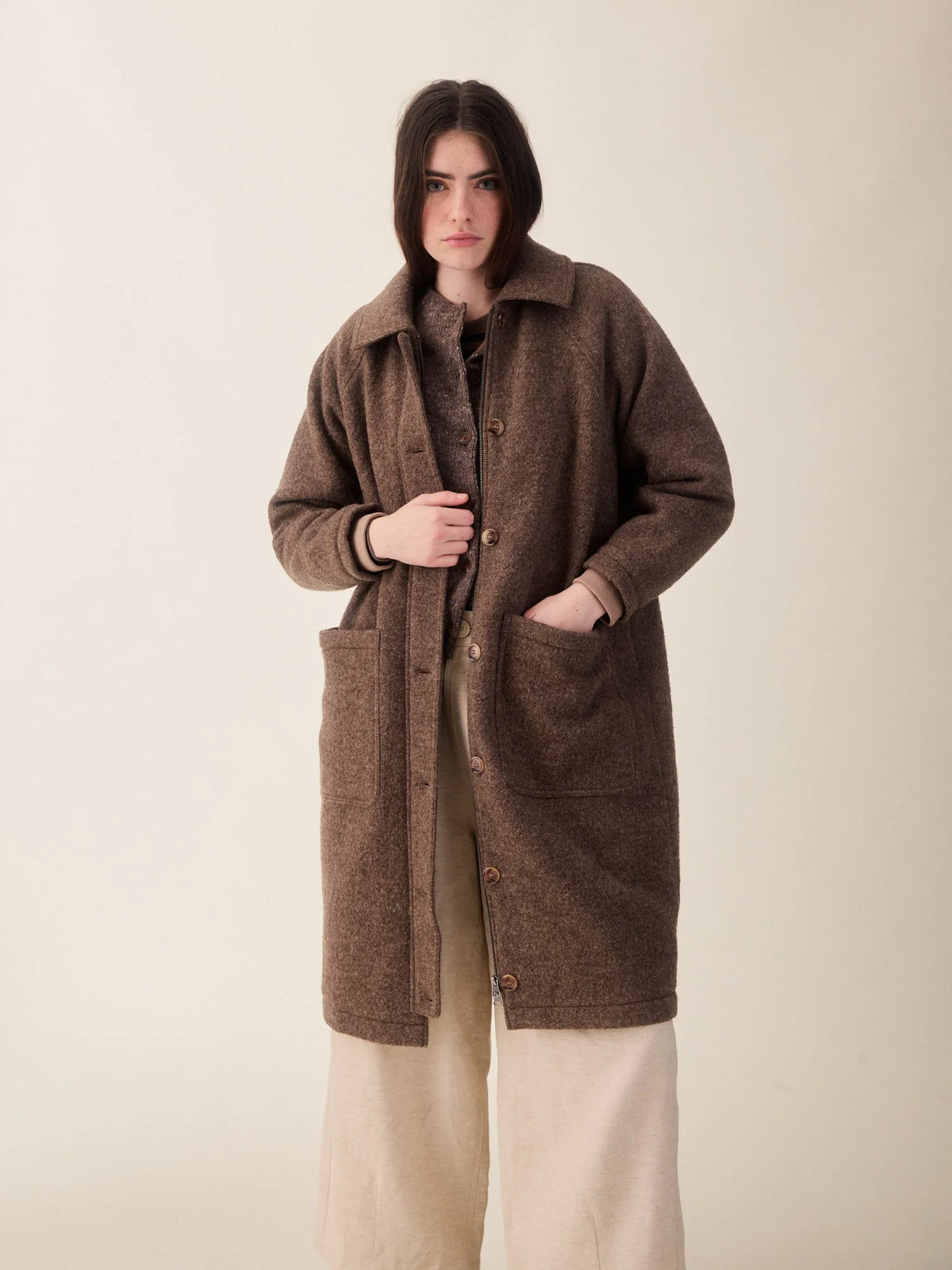 A woman wearing a brown textured mid-length coat with large front patch pockets over a beige outfit, standing against a plain light beige studio background.
