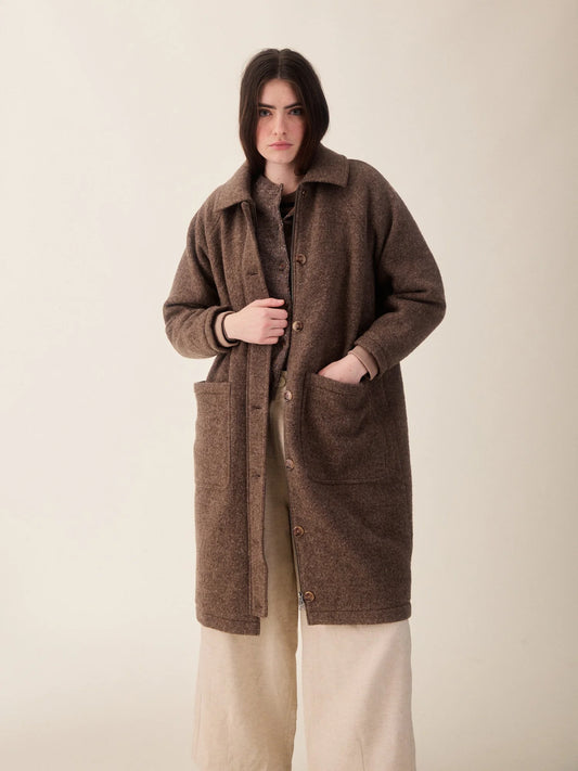 A woman wearing a brown textured mid-length coat with large front patch pockets over a beige outfit, standing against a plain light beige studio background.
