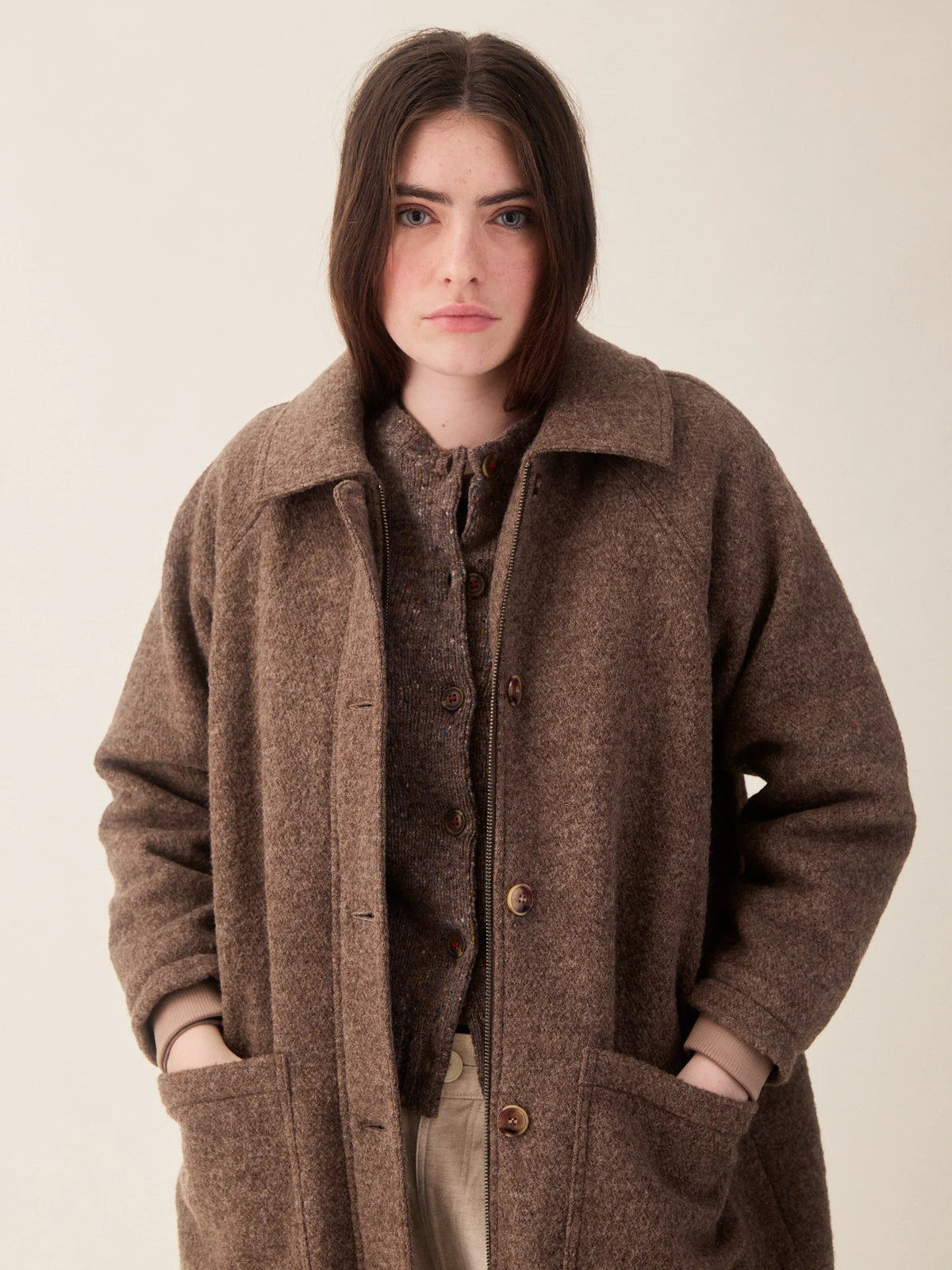 A woman wearing a brown textured wool-blend coat over a matching brown sweater and beige trousers, standing against a plain light beige studio background.