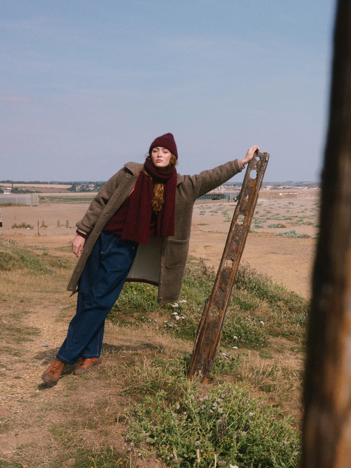 A person standing outdoors in a dry grassy field, wearing a brown coat, burgundy scarf and beanie, blue jeans, and brown boots, while leaning against a weathered fence.