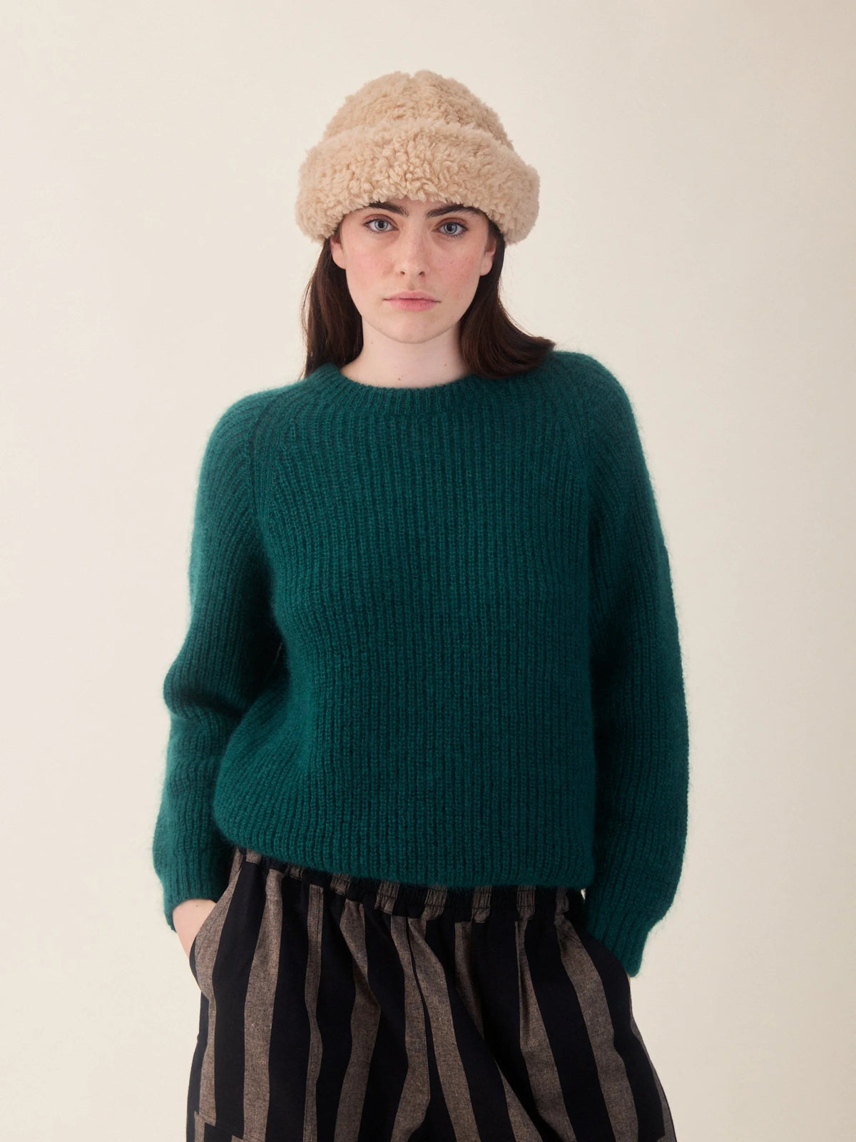 A model wearing a dark teal-green ribbed knit sweater, a beige fuzzy beanie, and black-and-brown striped trousers, standing against a plain light beige background.