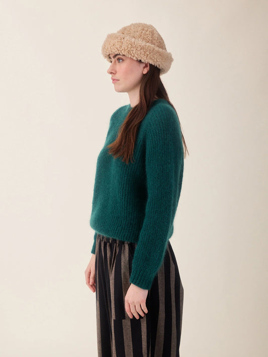 A woman in profile wearing a textured dark green knit sweater, a beige bouclé beanie, and dark striped trousers, photographed against a plain light beige studio background.