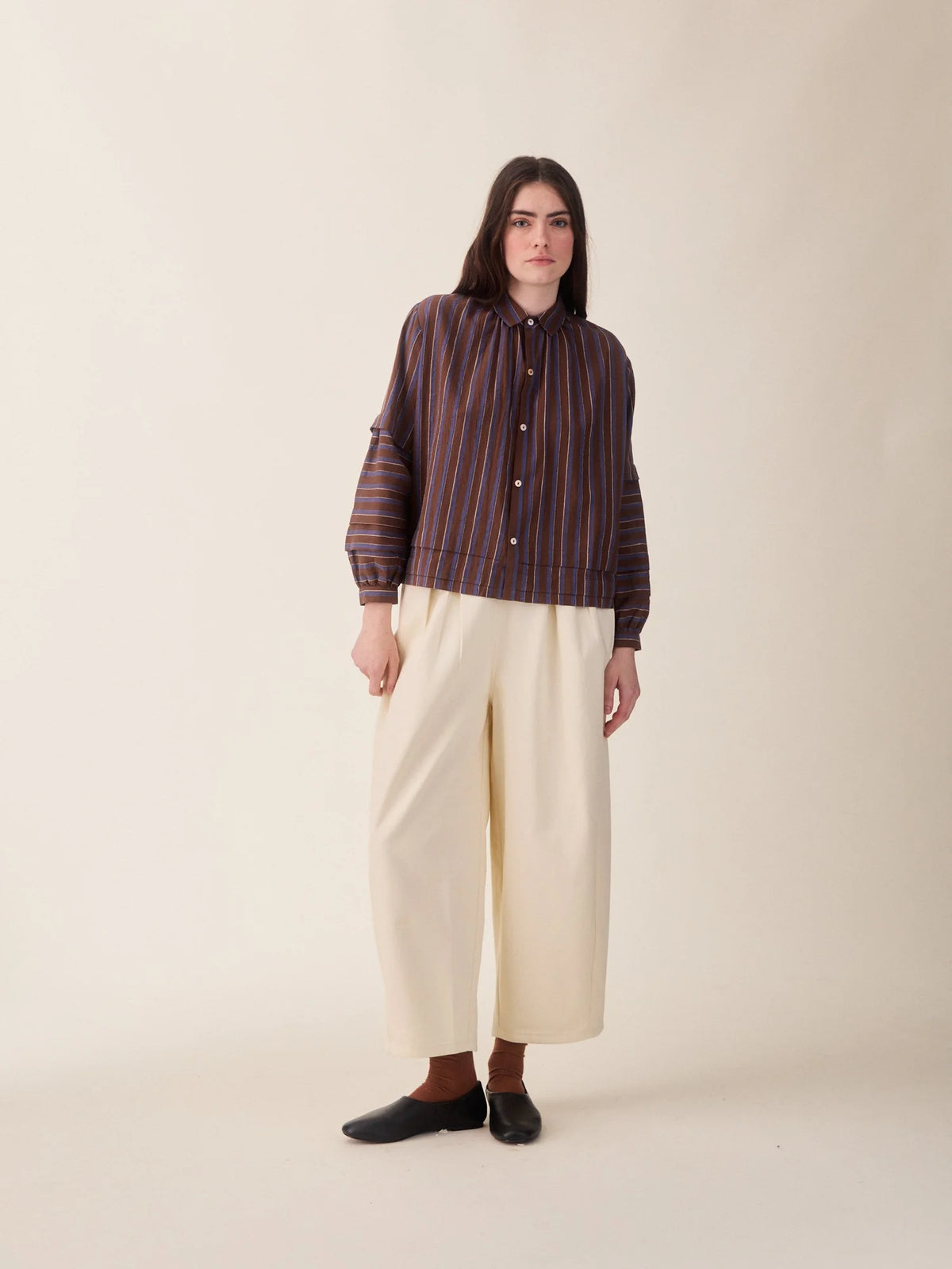 A model stands against a plain beige studio background wearing a dark brown and red vertically striped button-up shirt, cream wide-leg cropped pants, and black slip-on shoes.