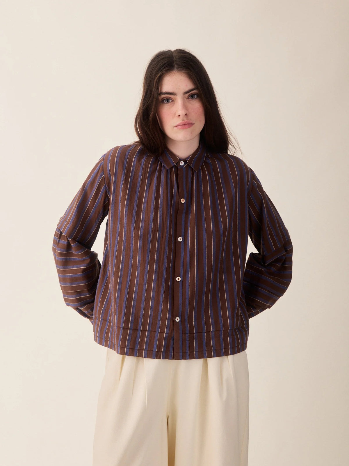 A woman modeling a dark brown button-up shirt jacket with vertical blue and white stripes, paired with cream-colored trousers, against a solid beige background.