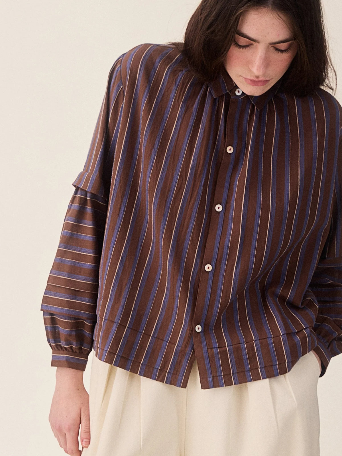 A woman wearing a brown button-up shirt with vertical blue and beige stripes, paired with cream-colored trousers, photographed against a solid light beige background.
