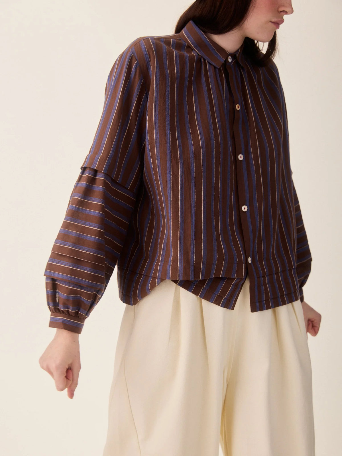 A model stands against a plain beige studio background wearing a dark brown and red vertically striped button-up shirt, cream wide-leg cropped pants.
