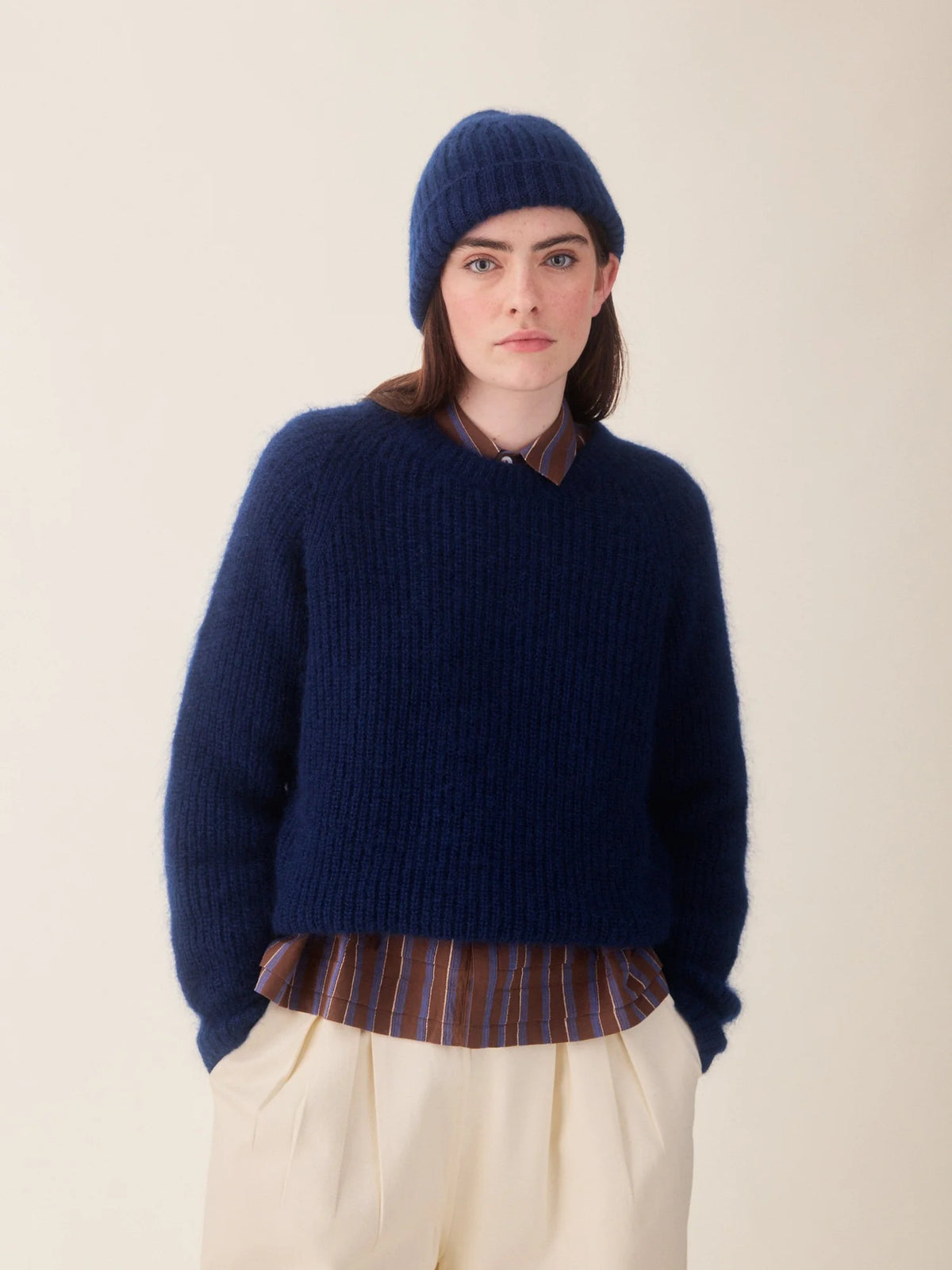 A model wearing a chunky navy blue knit sweater, matching navy beanie, cream trousers, and a striped shirt, photographed against a plain beige studio background.