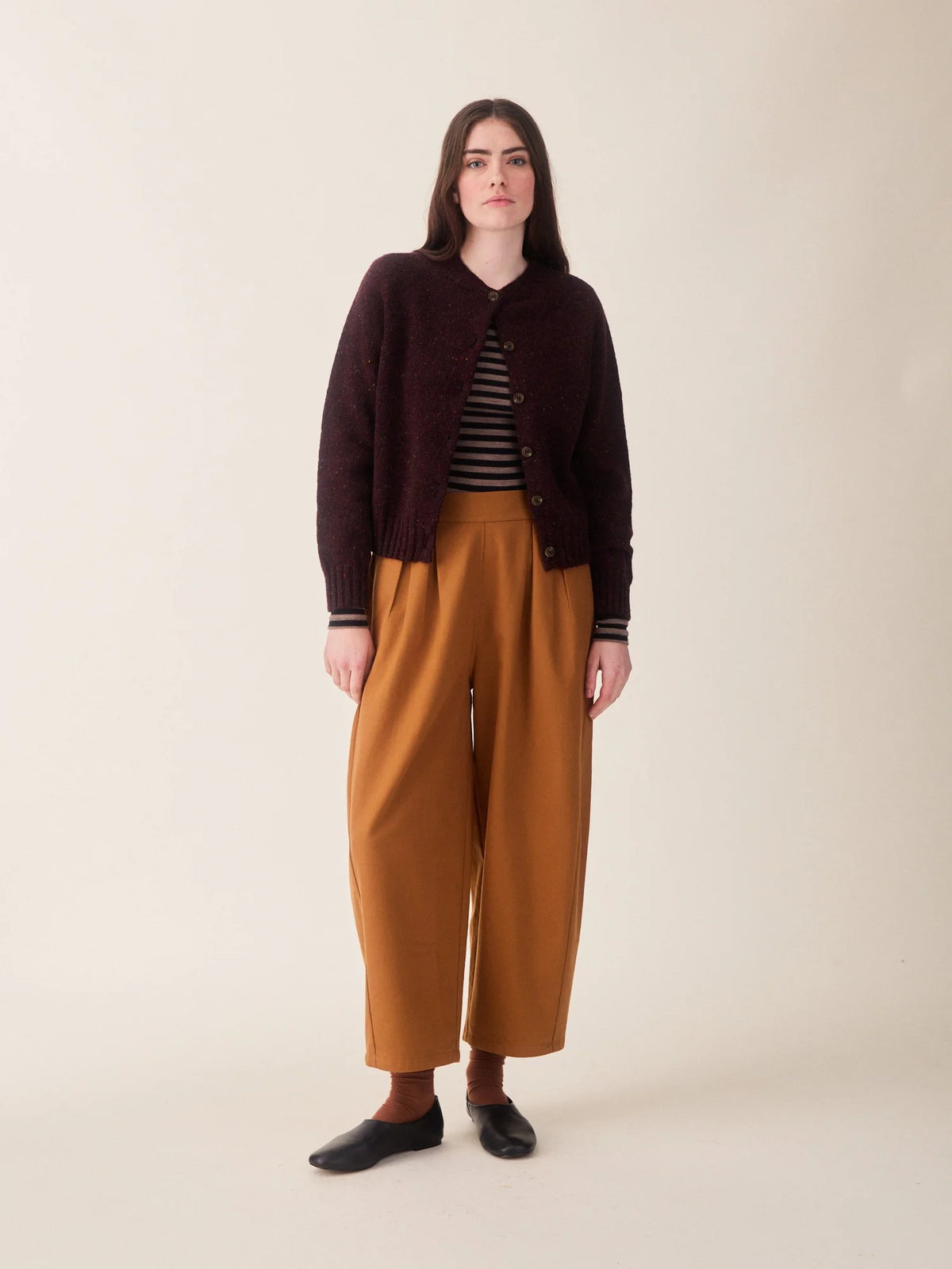 A woman wearing a dark burgundy heathered knit cardigan with brown buttons, layered over a black and beige striped top and paired with mustard yellow trousers, against a plain off-white background.