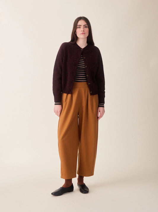 A woman wearing a dark burgundy heathered knit cardigan with brown buttons, layered over a black and beige striped top and paired with mustard yellow trousers, against a plain off-white background.