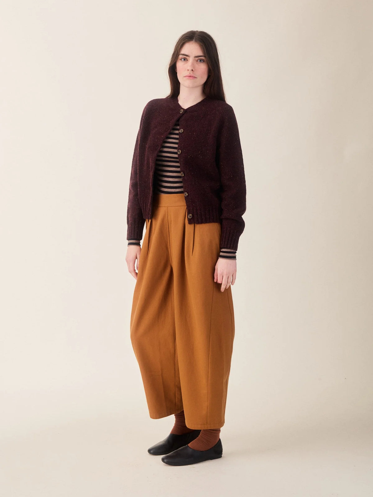 A model stands against a plain beige studio background wearing a dark burgundy knit cardigan over a black and white striped top, paired with wide-leg mustard-brown trousers, brown socks, and black slip-on shoes.