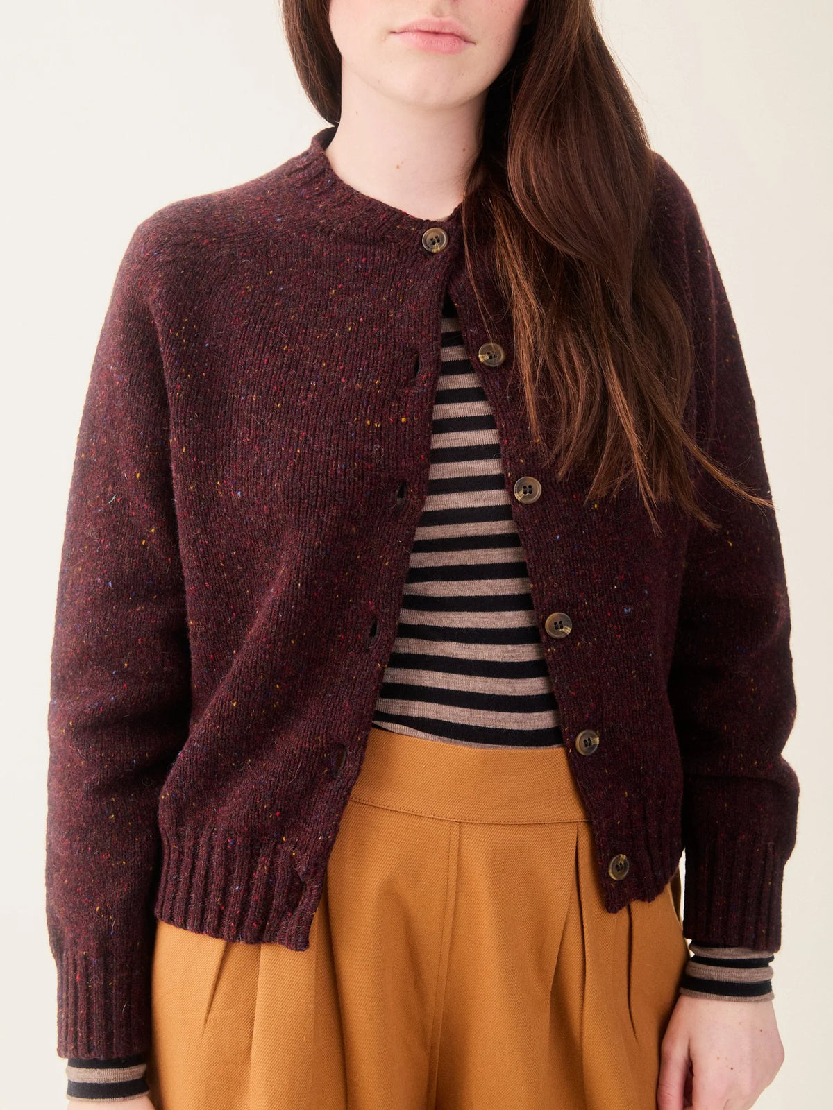 A woman wearing a dark burgundy heathered knit cardigan with brown buttons, layered over a black and beige striped top and paired with mustard yellow trousers, against a plain off-white background.