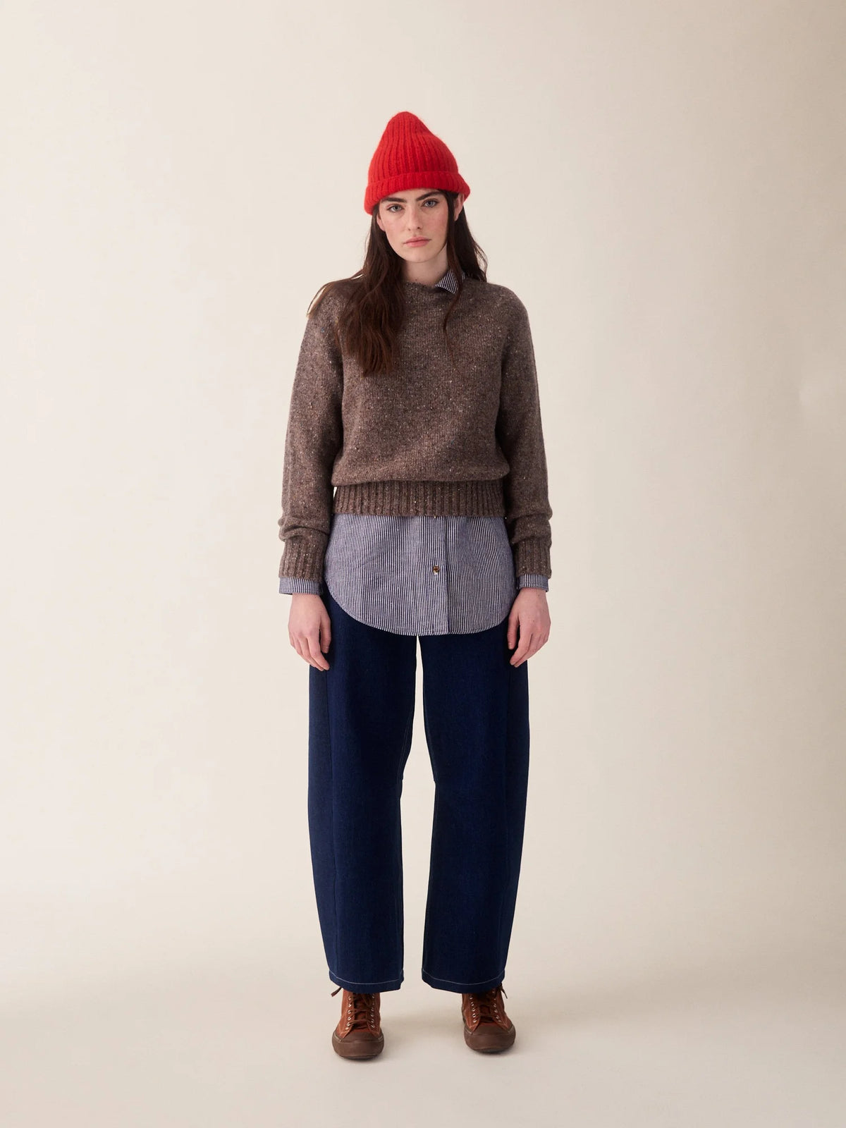 A model stands against a plain beige studio background wearing a brown knit sweater over a gray button-up shirt, dark navy wide-leg pants, and a bright red beanie.