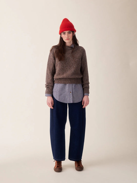 A model stands against a plain beige studio background wearing a brown knit sweater over a gray button-up shirt, dark navy wide-leg pants, and a bright red beanie.