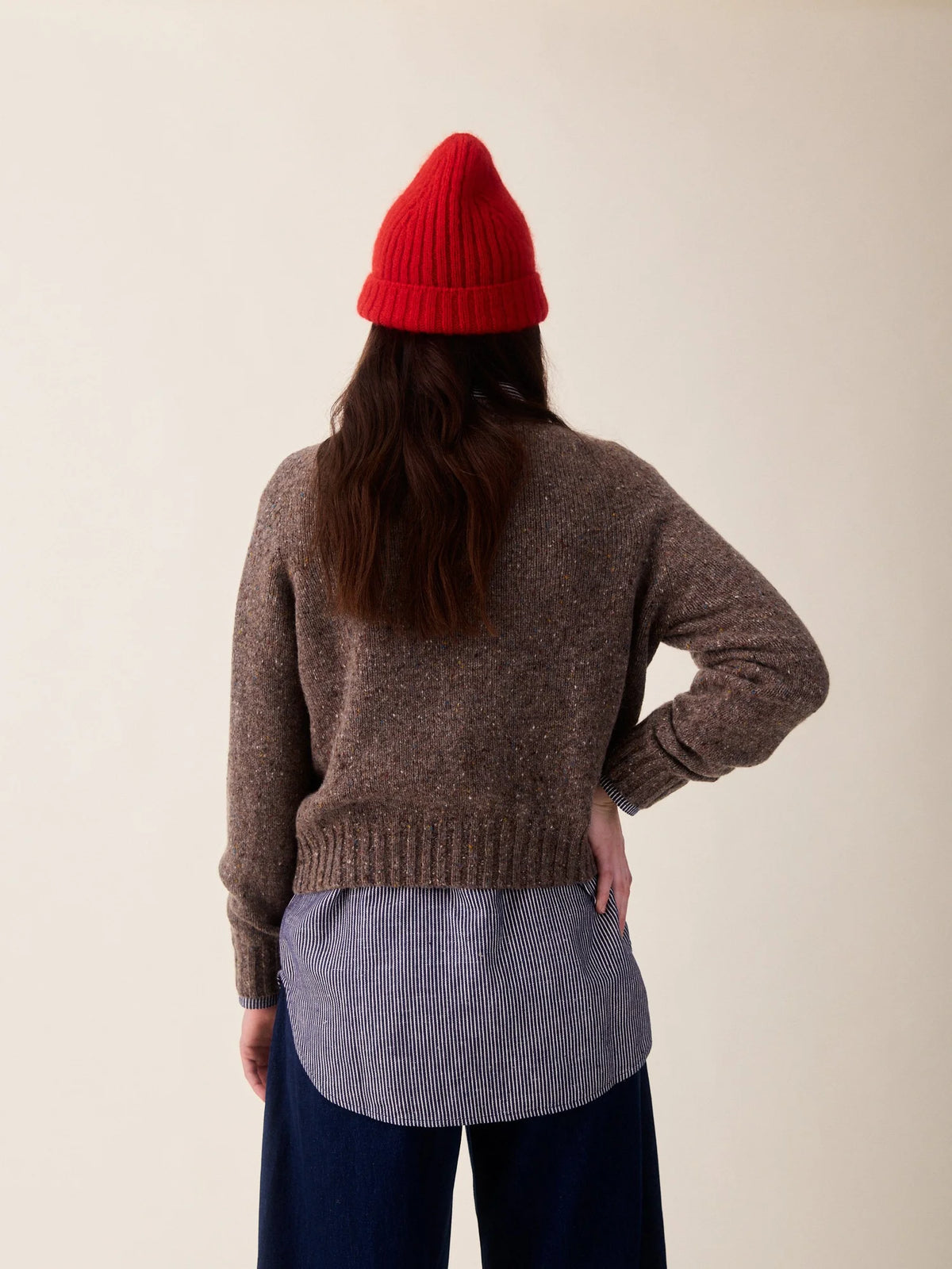 A person shown from the back wearing a brown knit sweater, a red beanie, a light blue striped shirt, and dark blue pants against a plain beige studio background.