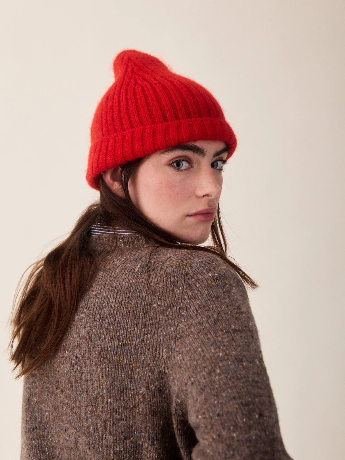 A woman wearing a bright red ribbed knit beanie and a brown tweed sweater, photographed against a plain light beige studio background.
