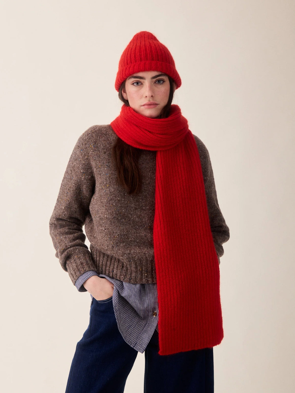 A model wearing a brown knit sweater, a bright red ribbed scarf, a matching red beanie, and dark blue pants, standing against a plain light beige background.